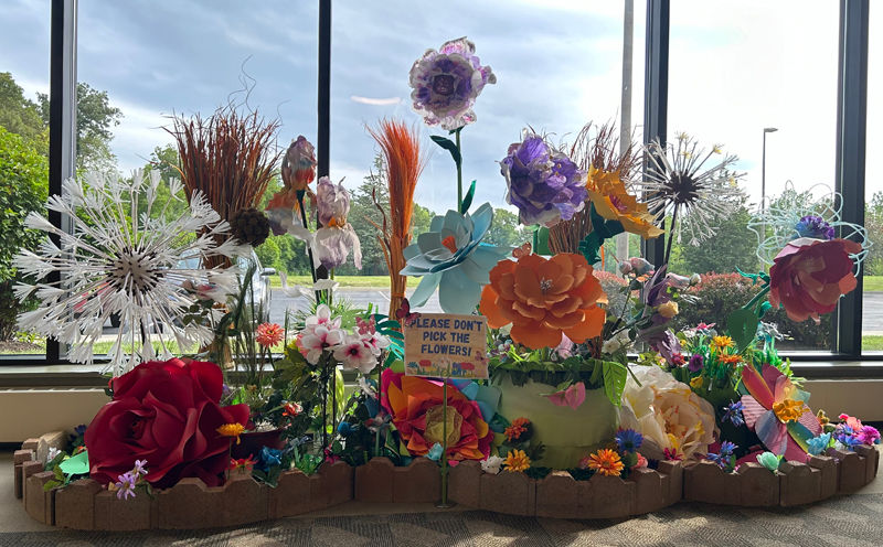 Colorful paper flowers of various shapes and sizes displayed indoors near a large window, with a sign reading ‘Please don’t pick the flowers!’