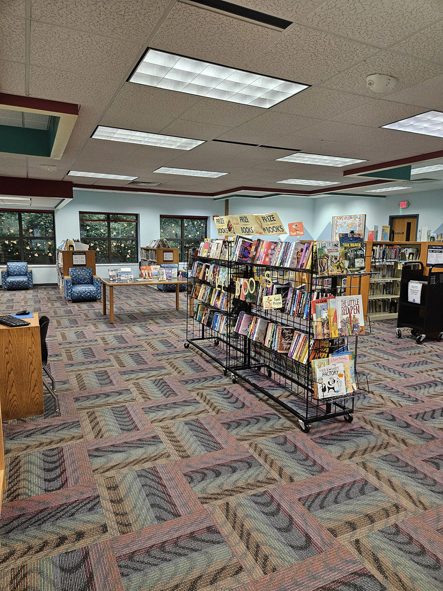 Interior of the Aboite Branch library with colorful carpet, bookshelves filled with books, bright overhead lights, and large windows.