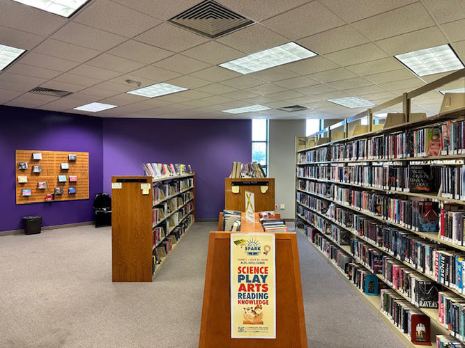 Library interior with rows of bookshelves near the teen area, and a colorful poster promoting science, play, arts, and reading.