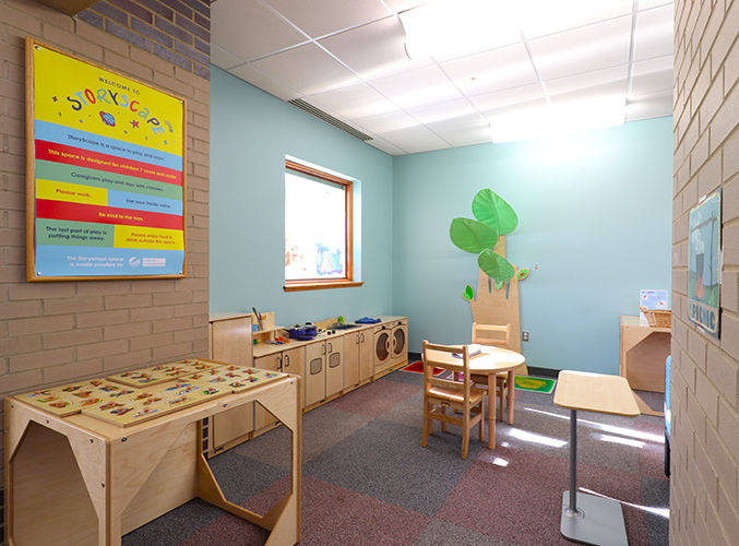Children’s Storyscape area with wooden furniture, a wooden play kitchen, a ‘Story Scape’ wall poster, a child-sized table and chairs, and a large interactive leaf and tree decoration.