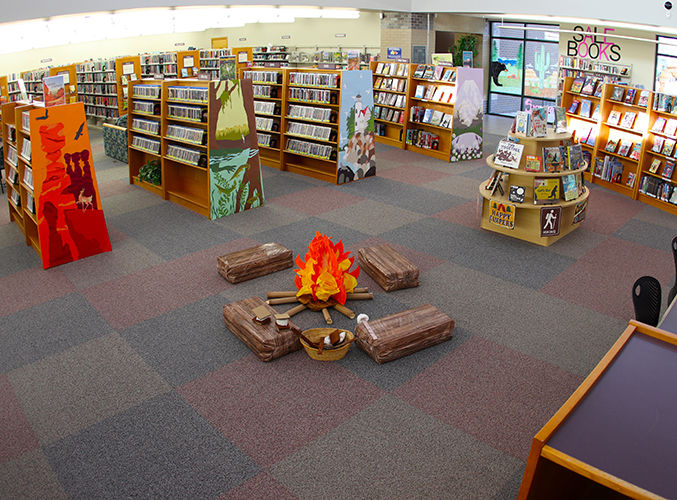 Library interior with wooden bookshelves and vibrant end-of-shelf artwork, featuring a central play campfire display with cushioned fake logs for children to sit on.