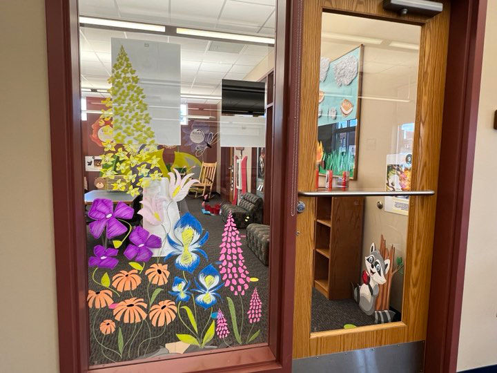 Glass door with colorful paper flowers and foliage painted on the window. Inside is a room with books, chairs, and a wall decorated with artwork.