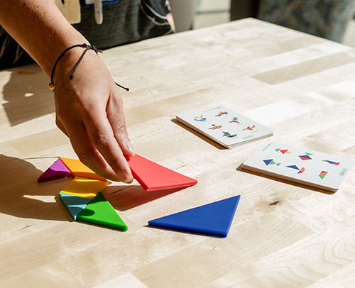 Hand arranges colorful tangram puzzle pieces on a table beside challenge cards.