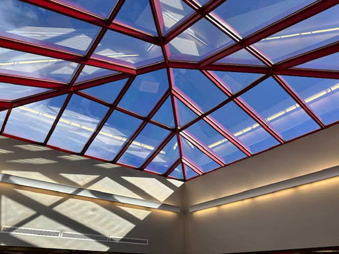Glass ceiling with a red triangular grid pattern casting geometric shadows on the wall, with bright blue sky visible through the glass.