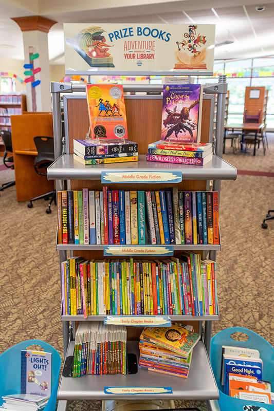 A library book shelf labeled Prize Books Adventure Your Library displays children's and middle-grade fiction.
