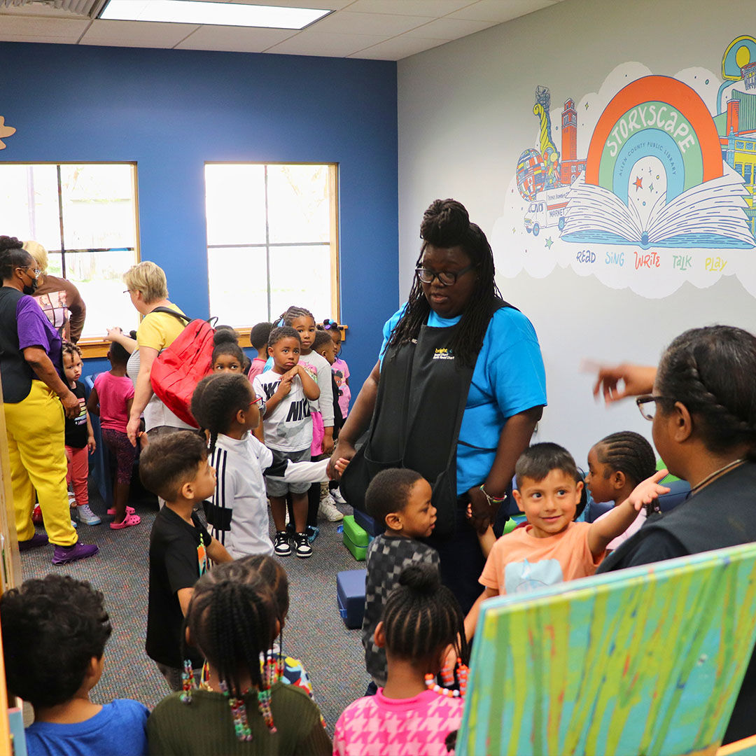 A group of young children and caregivers gather in a Storyscape area with blue walls and a mural of an open book with the Storyscape logo.