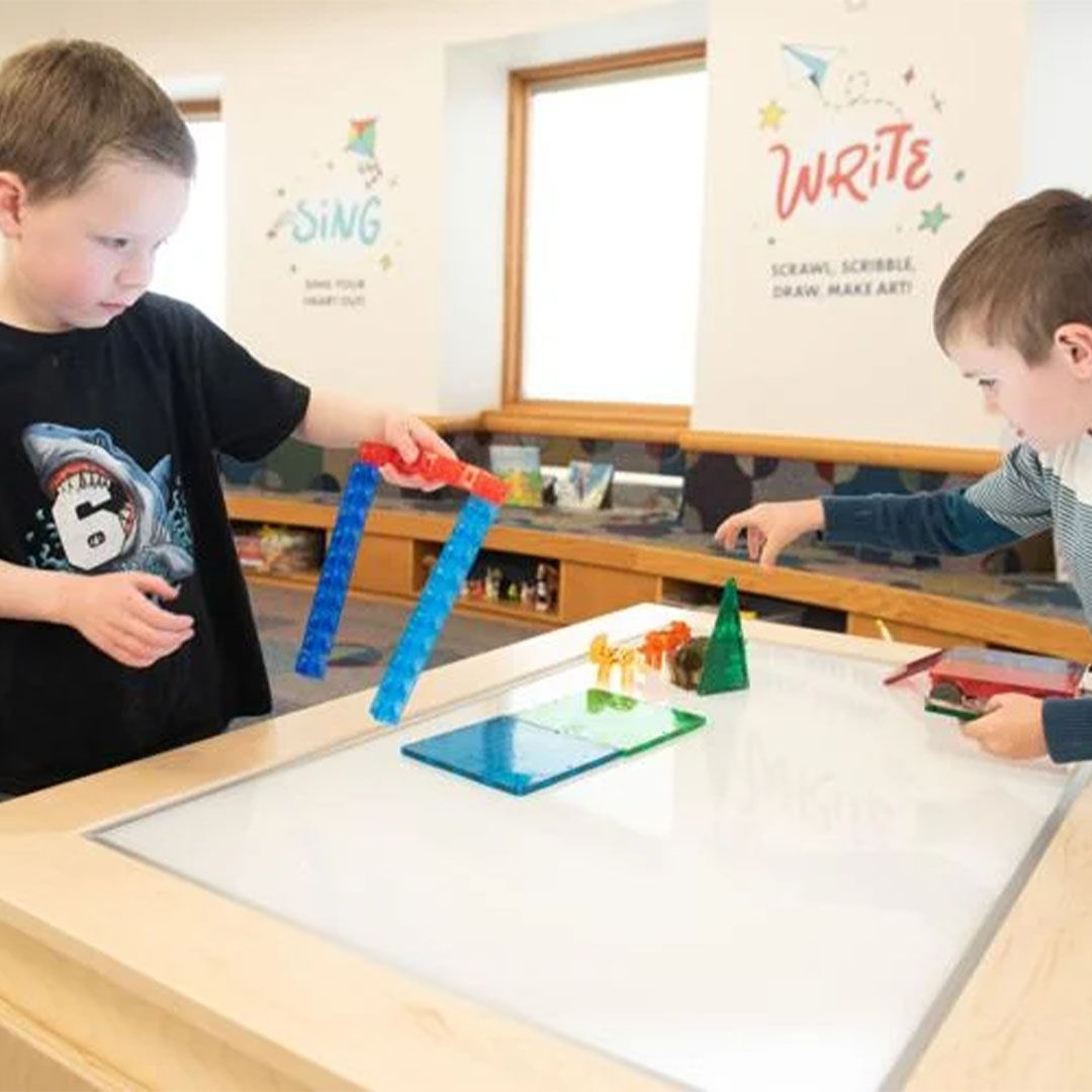 Two young boys playing at a light table with colorful magnetic tiles, with playful wall art in the background featuring the words Sing and Write.