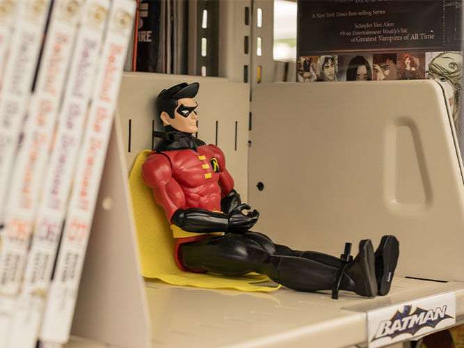 Figure of Robin in red and black sitting on a bookshelf with crossed arms, surrounded by comic books and a ‘Batman’ label.