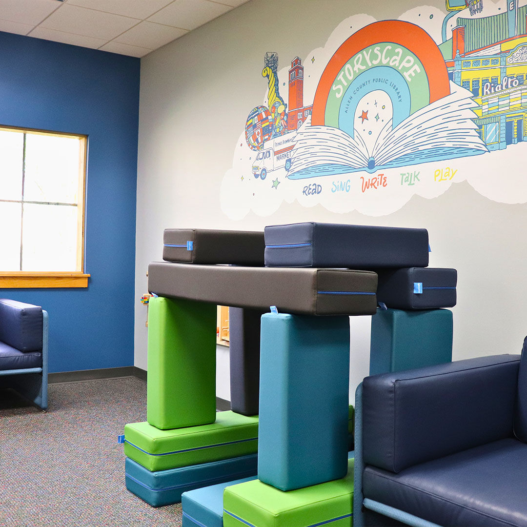 Storyscape area with colorful foam blocks and seating. A Storyscape logo mural on the wall features an open book and the words Read Sing Write Talk Play.