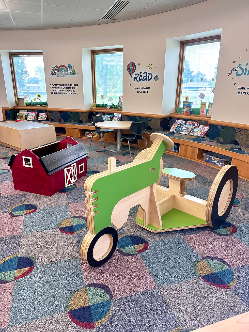 StoryScape area at Woodburn Branch with a climbable green wooden tractor, red barn toy, and curved wall seating with cubbies of books and toys.