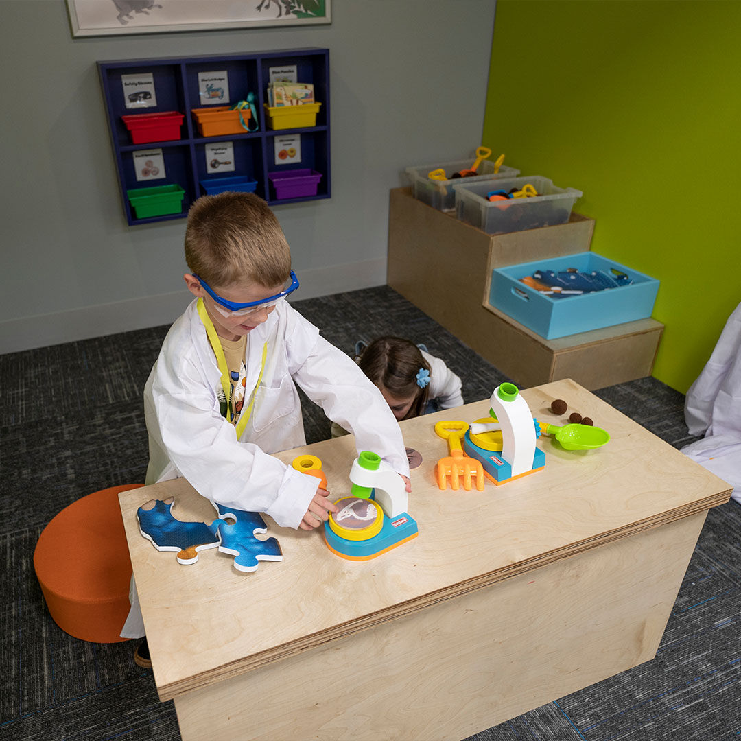 A child wearing a lab coat and goggles playing with toy science equipment at a wooden table, with colorful bins and lab tools in the background.