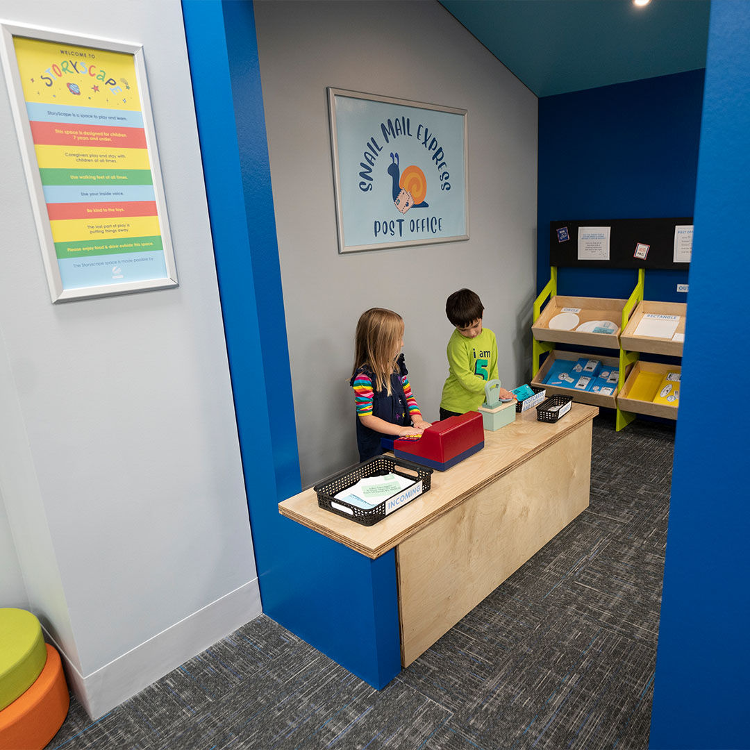 Children play in a colorful, kid-sized post office with vibrant walls, engaging with a counter and mail items.