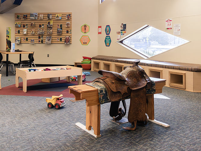 Library children’s area with a saddle on a wooden stand, toy truck, activity table, colorful wall art, and a diamond-shaped window.