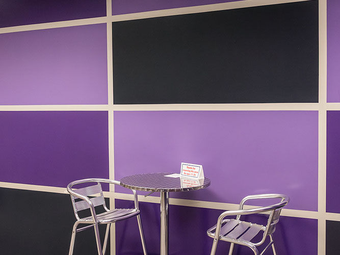 Two metal chairs and a round table are positioned against a purple and black checkered wall in the teen area.