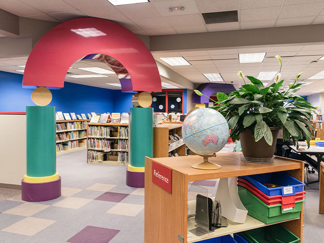Library interior of the children’s area with a colorful archway, bookshelves filled with books, and a globe on a reference desk beside a large leafy plant.