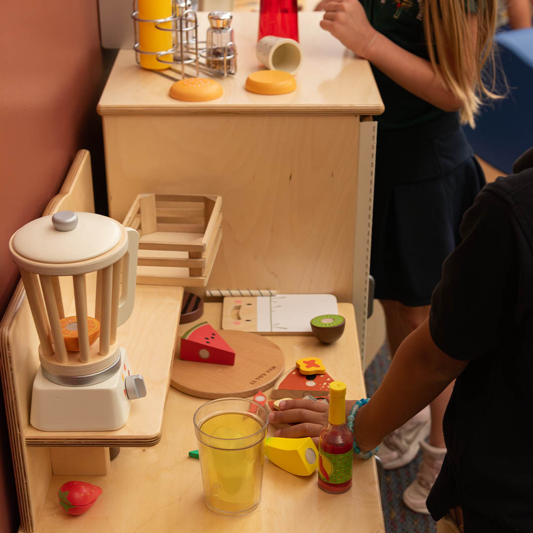 Children playing with a wooden toy kitchen set, including a toy blender, fruit, and condiments.