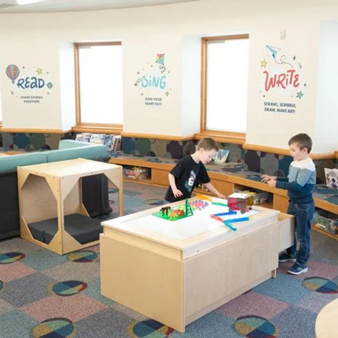 Children play with toys on a light table in a Storyscape area, with colorful wall signs encouraging reading, singing, and writing.