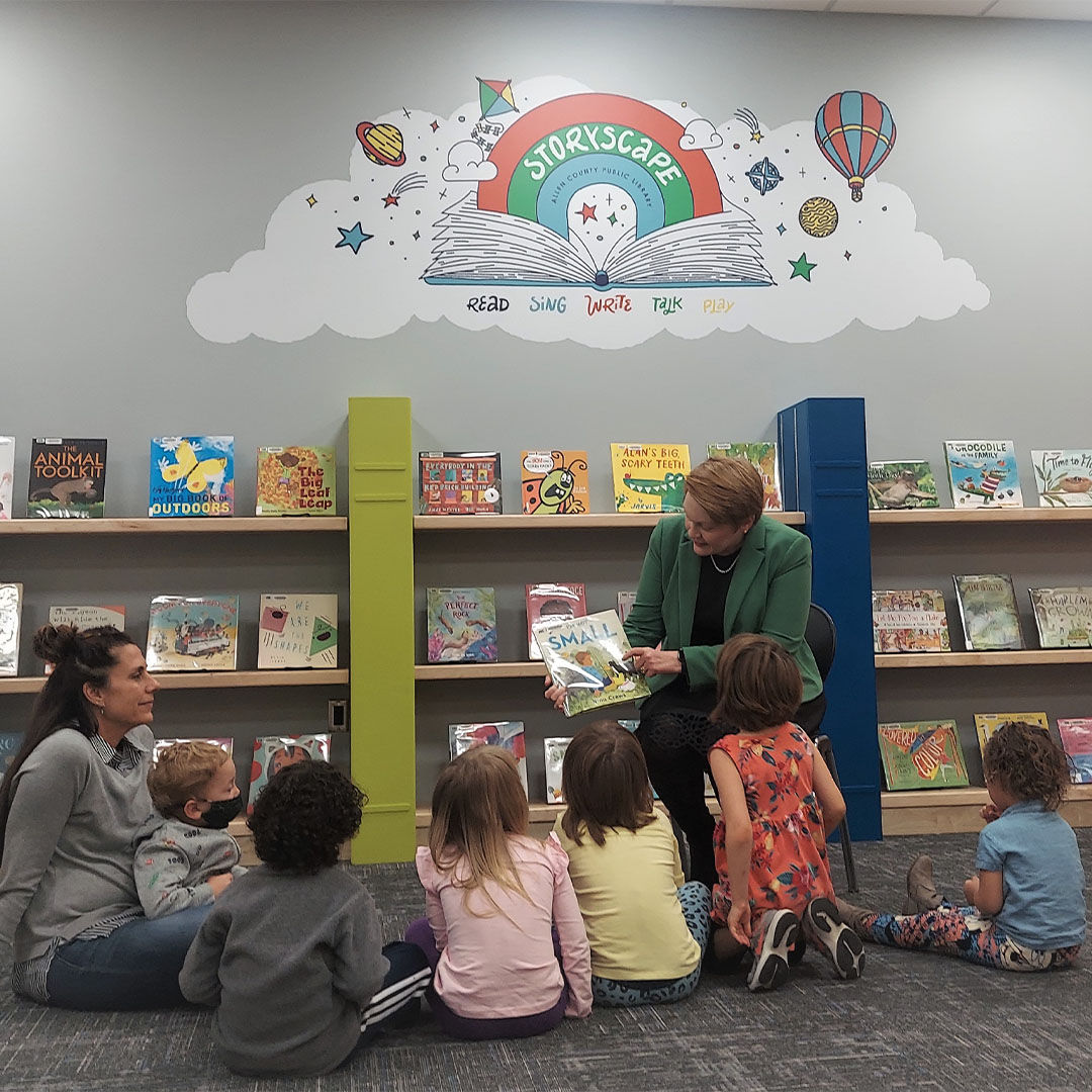 A woman reads to a group of young children seated on the floor in a Storyscape area of the library, with shelves of colorful books and a Storyscape mural in the background.