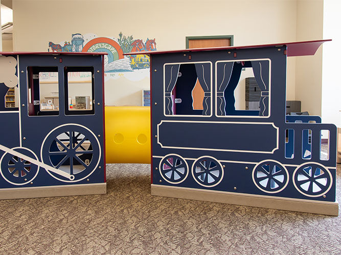Children's play area featuring a blue train structure with white outlines and windows. A yellow tunnel connects the cars.