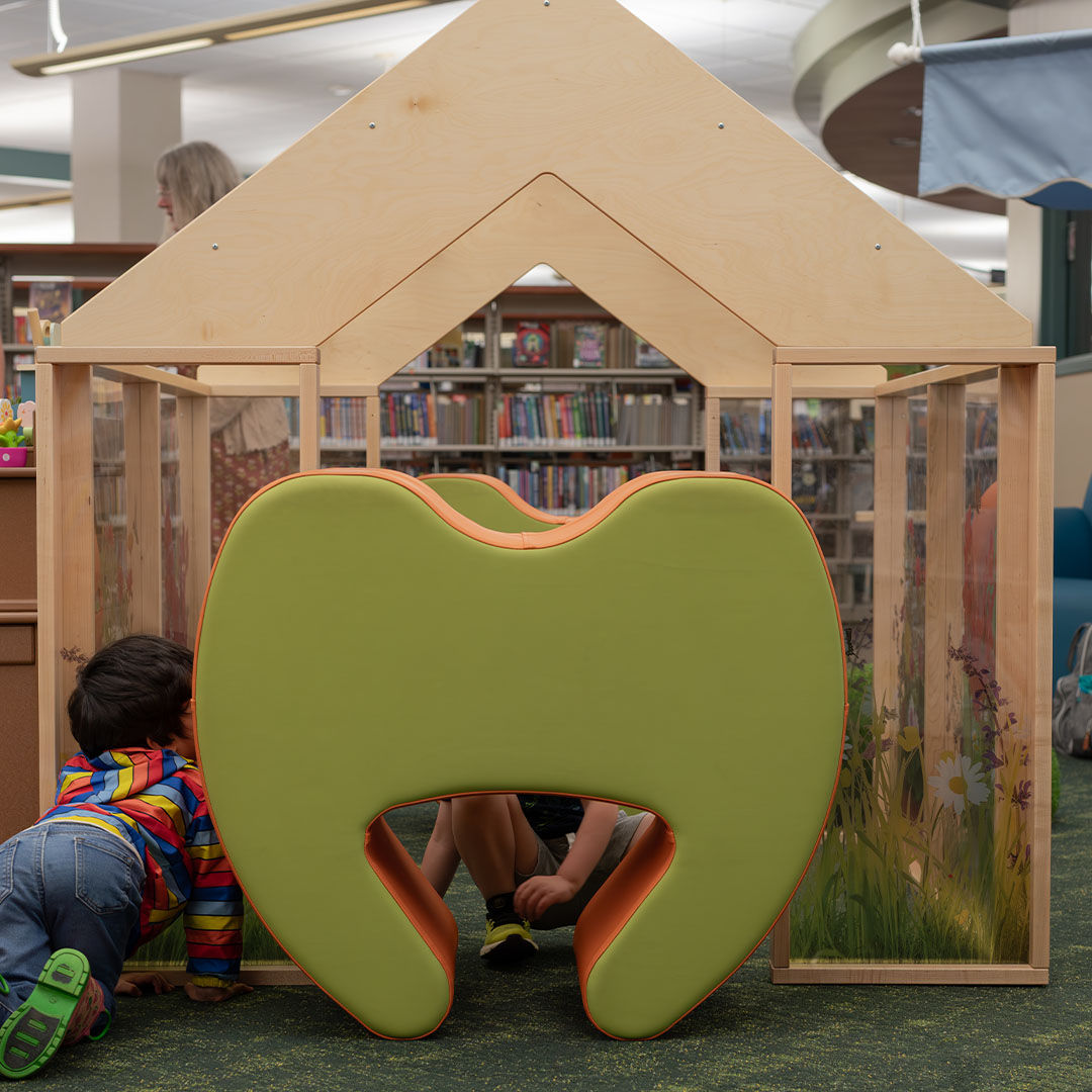 Children play inside a wooden greenhouse structure with a green and orange cushioned seat at its opening, with shelves of books visible in the background.