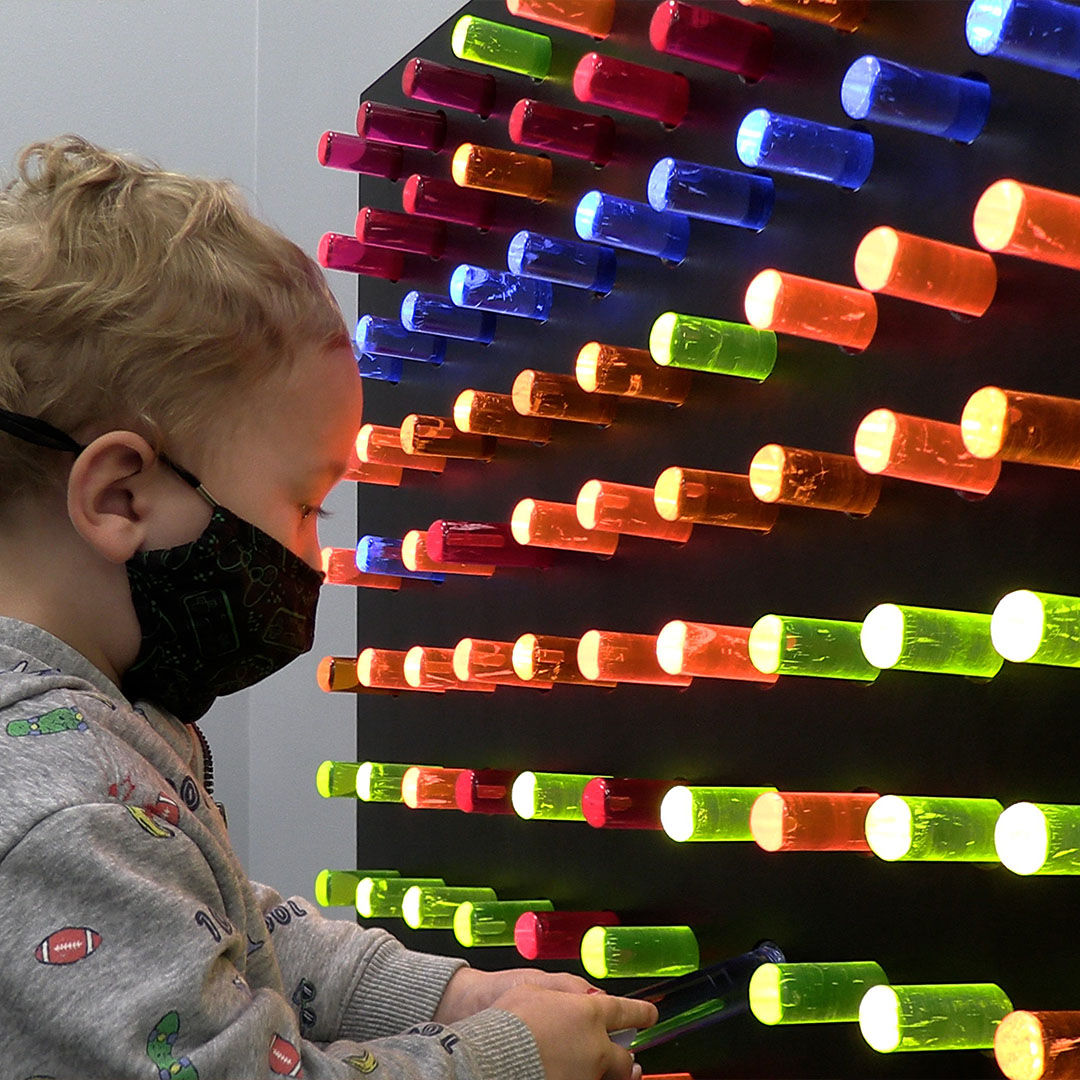 A young child wearing a mask interacts with a colorful pegboard wall, with pegs glowing in red, blue, green, and orange.