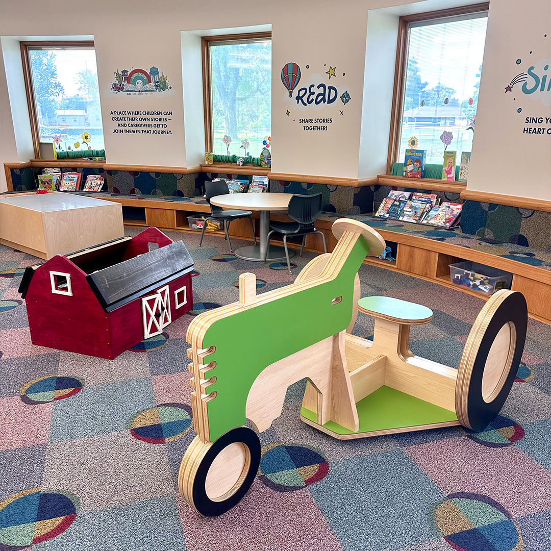 Children’s Storyscape area with a wooden climb-on green tractor and red toy barn surrounded by books on shelves and colorful carpet patterns.