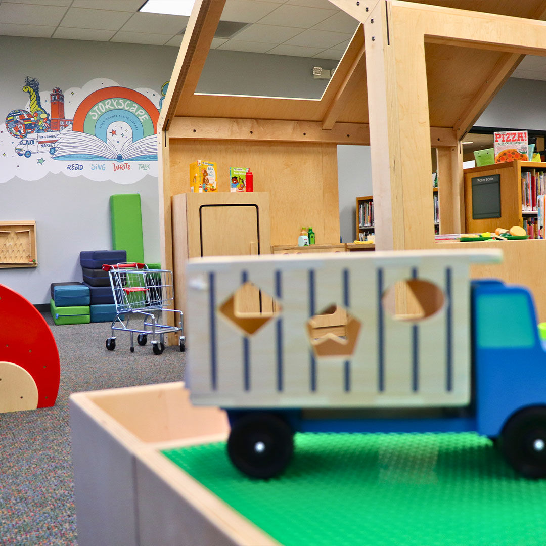 Storyscape area featuring a toy truck, a small shopping cart, and colorful cushions, with a wall mural reading Storyscape and a wooden structure in the background.