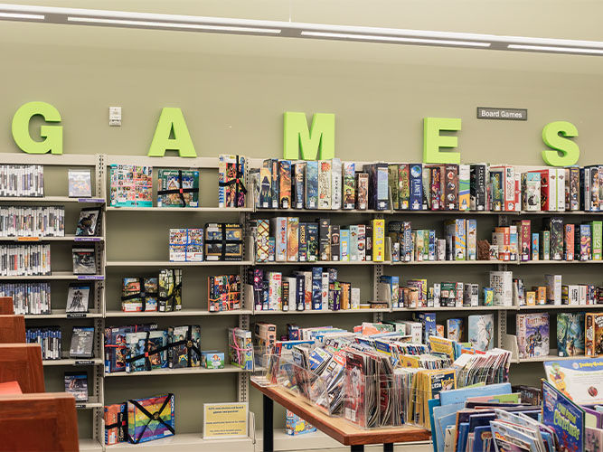 Library section with shelves full of board games under large green letters spelling ‘GAMES,’ with transparent bins of magazines in front.