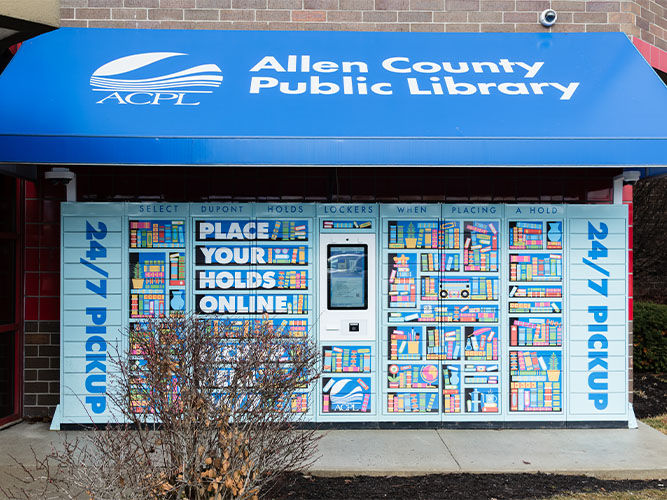 Outdoor library book locker with vibrant bookshelf design, covered by a blue awning labeled Allen County Public Library. Text reads 24/7 Pickup.