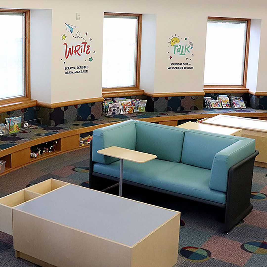 A Storyscape area with colorful cushioned benches for children, a small teal couch in front of a table, and bookshelves filled with books.