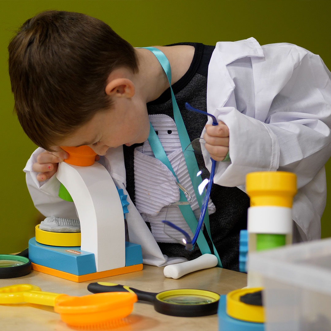 A young child in a white lab coat peers through a toy microscope on a wooden table, with brightly colored magnifying glasses and tools surrounding him.