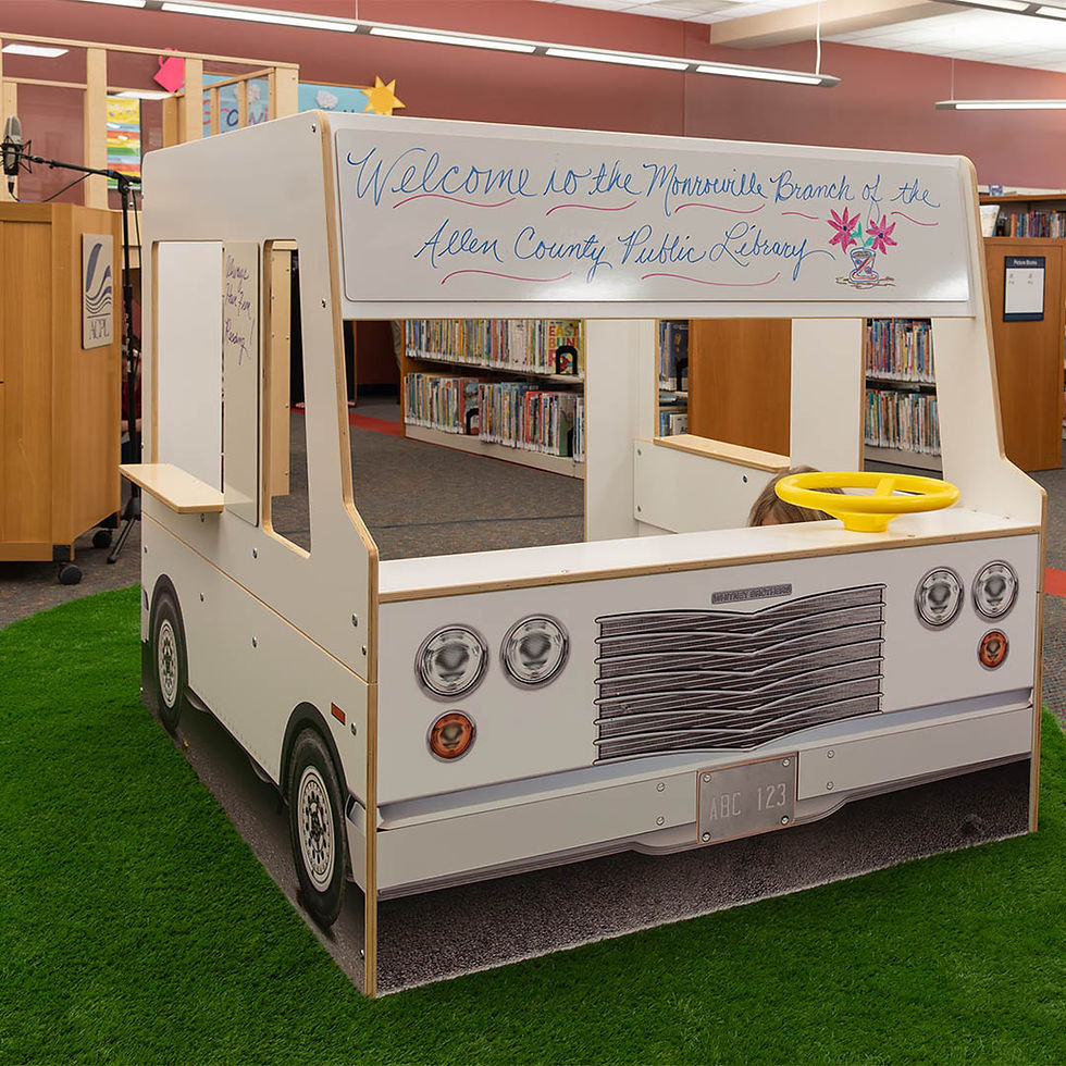 White interactive food truck with a yellow steering wheel at the Monroeville StoryScape area on green carpet. Sign reads ‘Welcome to the Monroeville Branch of the Allen County Public Library,’ with bookshelves behind.