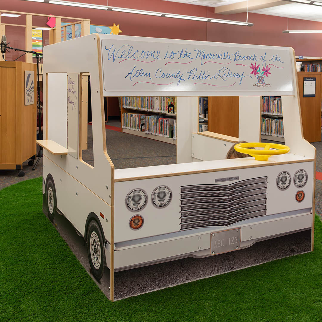 Wooden play structure resembling a food truck inside a library with a welcome message on top, set on green carpet with colorful bookshelves in the background.