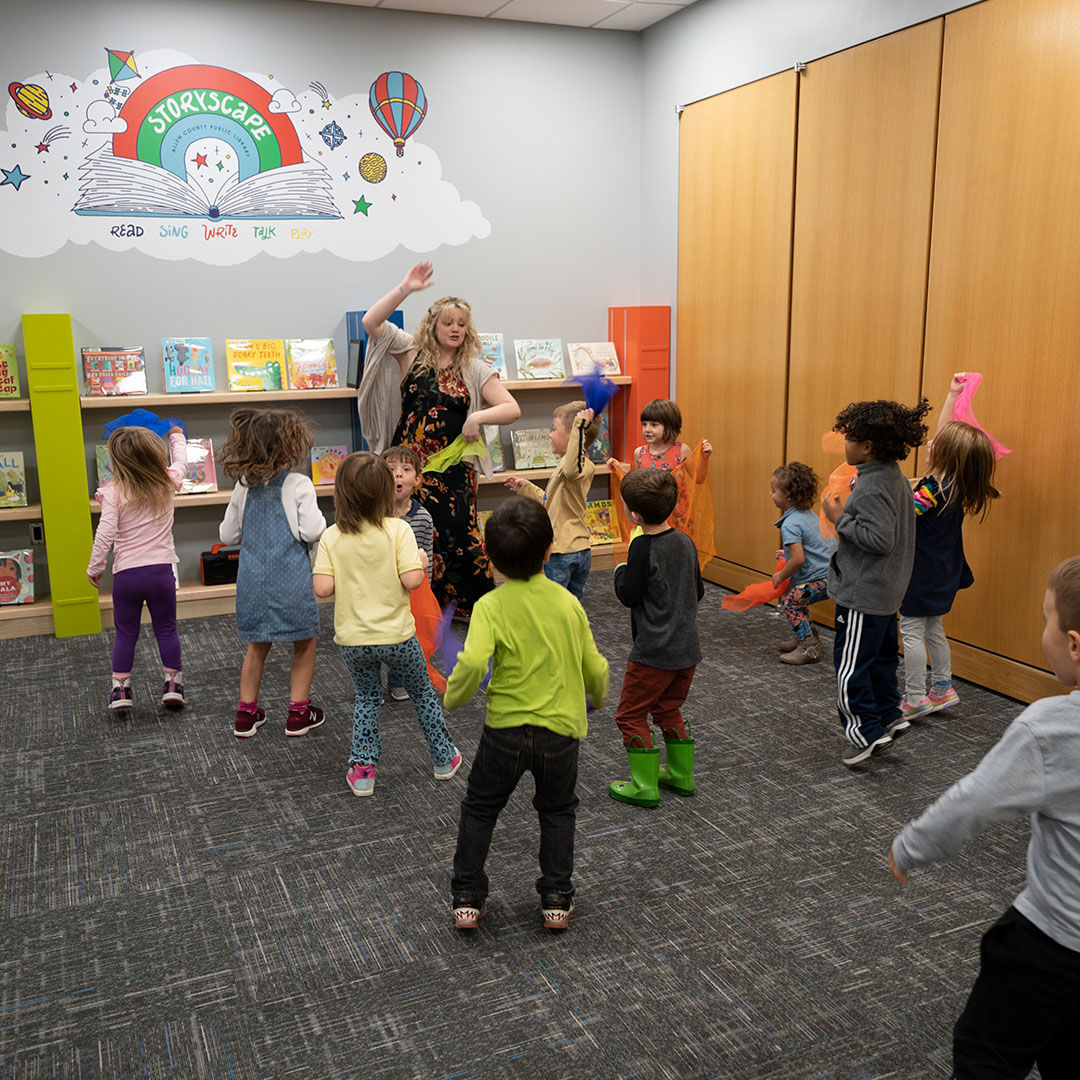 Storytime at the library with a group of children dancing joyfully around a librarian, with books lining shelves in the background under a Storytime mural.
