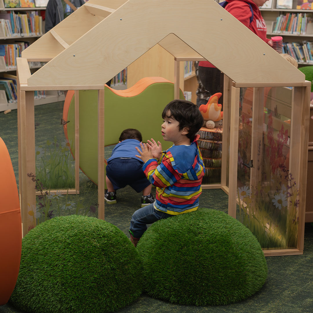 Two children play in the Storyscape area. One sits on round, grass-like cushions near a wooden greenhouse, while the other crawls through the greenhouse.