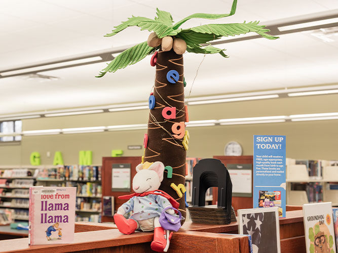 Plush tree with alphabet letters and a stuffed llama on a library bookshelf, alongside children’s books and a ‘Sign Up Today’ notice.
