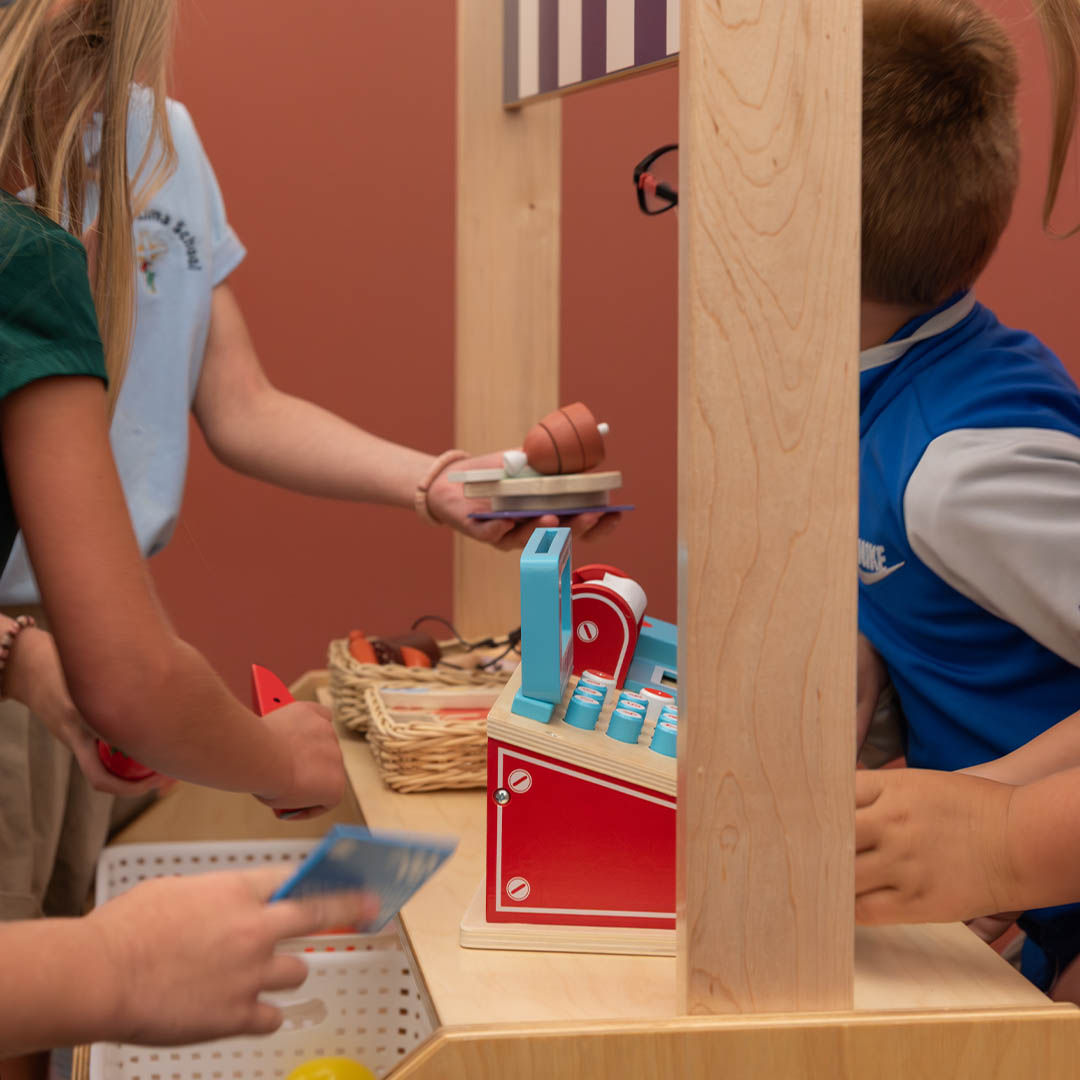 Children play with a toy cash register in a pretend market setup, holding play food items and cards.