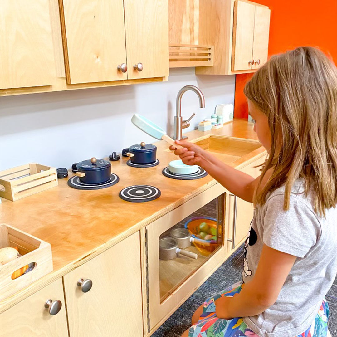 A young child playing in a wooden toy kitchen with blue pots and a spatula.