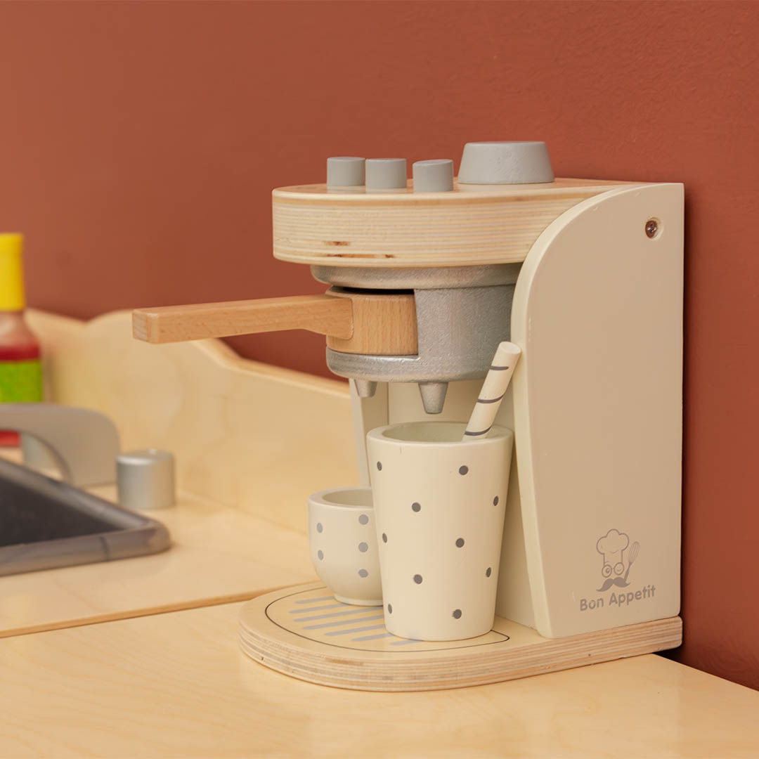 Wooden toy espresso coffee maker set against a brown wall with two spotted cups and a striped straw.