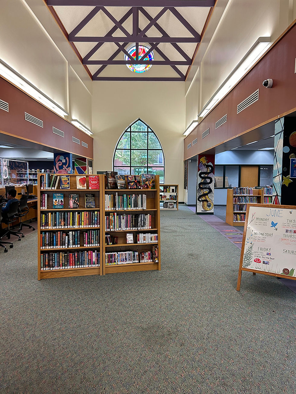 Interior de la biblioteca con techos altos abovedados, una estantería central con libros variados, un caballete con un letrero a la derecha, una zona de ordenadores a la izquierda y un gran ventanal arqueado con una vidriera circular encima, al fondo.