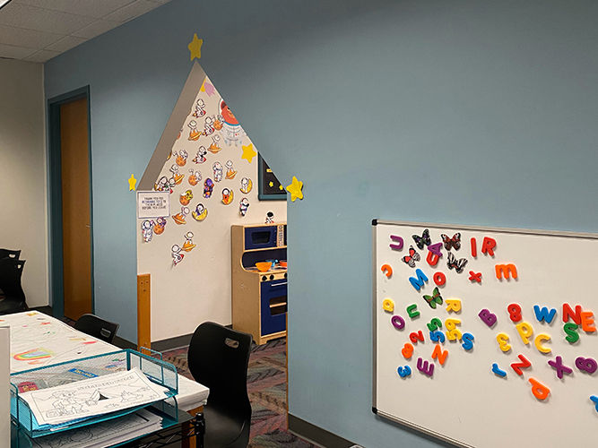 Una pared azul con un tablero magnético cubierto de imanes con letras del alfabeto y un arco con forma de casa que da acceso a la zona de juegos infantiles.