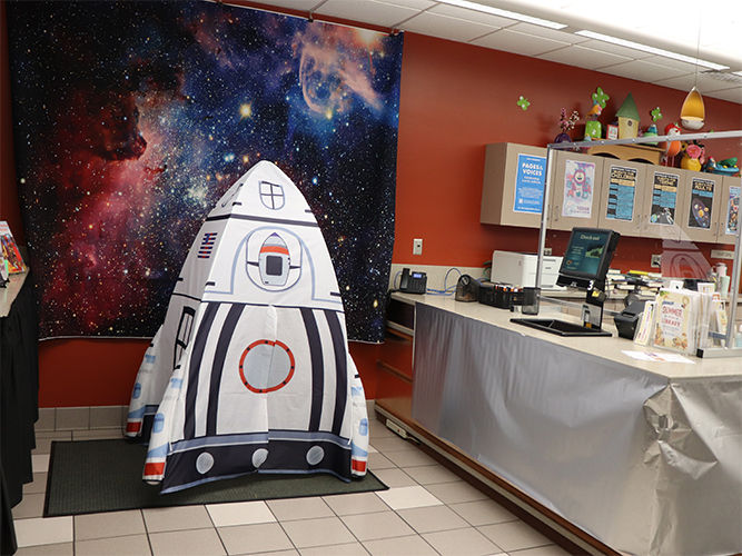 Pequeña réplica de una nave espacial en una biblioteca, con un fondo de temática galáctica y una zona de autopréstamo cerca.