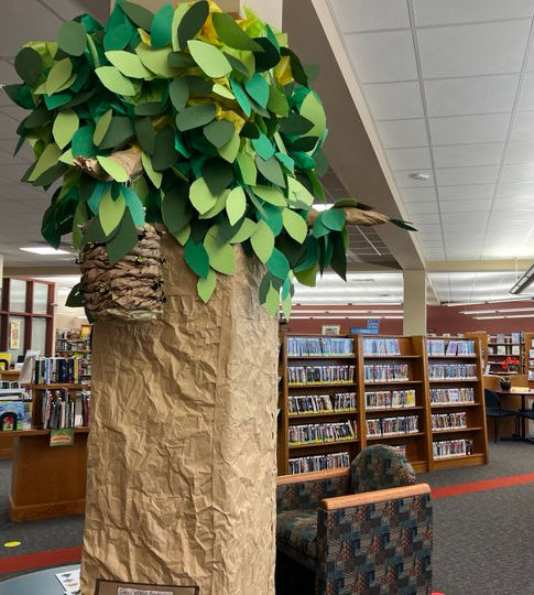 Árbol artesanal con tronco de papel arrugado y hojas verdes en una biblioteca, con estanterías en la pared y una silla cómoda cerca.