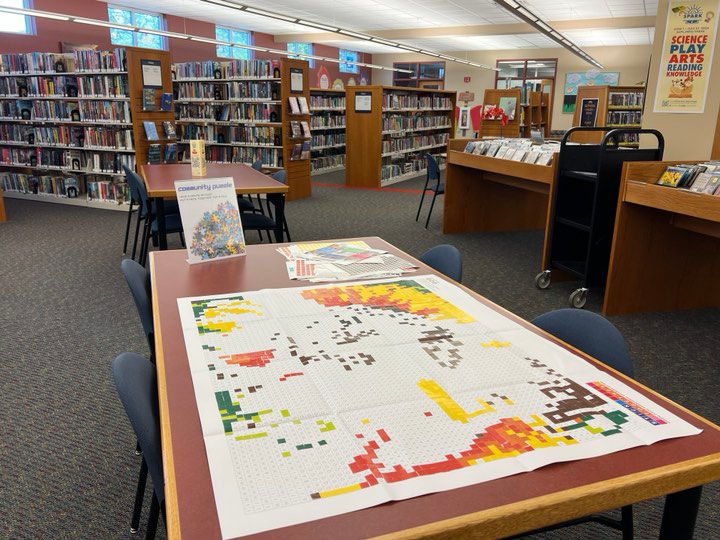 Mesa con un rompecabezas de mosaico incompleto compuesto por secciones coloridas, ambientada en el interior de una biblioteca con estanterías detrás.