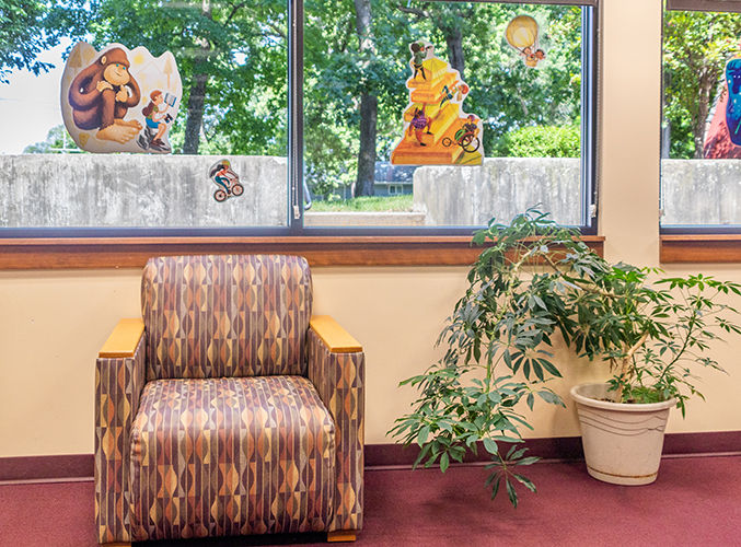 Un rincón de biblioteca con un sillón estampado junto a una planta frondosa en maceta, y ventanas decoradas con coloridas ilustraciones de animales y motociclistas.
