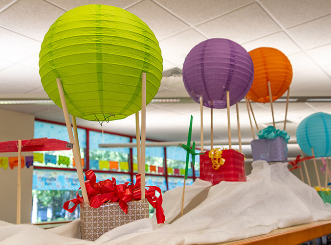 Farolillos de papel de colores, que recuerdan a globos aerostáticos, flotan sobre cajas de regalo envueltas en papel, colocadas sobre una superficie blanca cubierta con un drapeado.