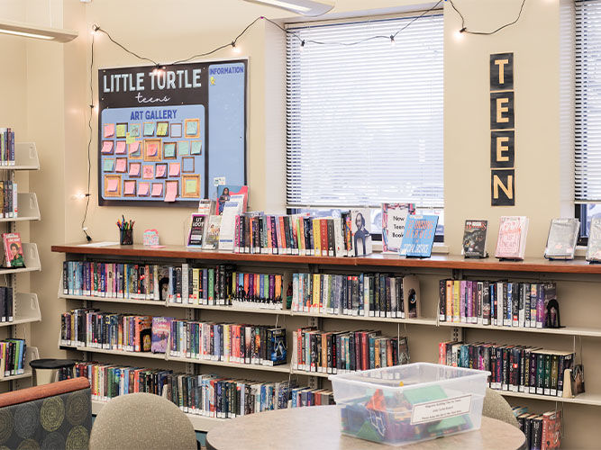 Sección juvenil de la biblioteca con estanterías llenas de libros coloridos, un tablón de anuncios con notas adhesivas, guirnaldas de luces y un letrero en la pared que dice "Adolescentes".