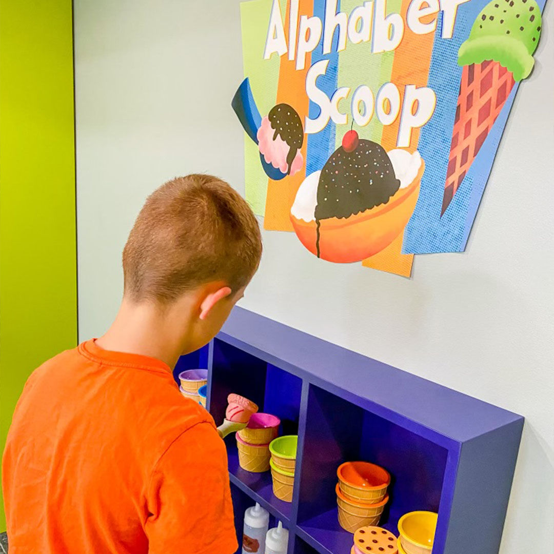 A child in an orange shirt interacts with a colorful educational display titled Alphabet Scoop, with play ice cream cones and bowls.