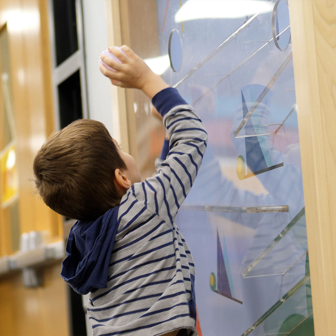 A young child in a striped shirt plays with an interactive wall toy, watching as a ball rolls down colorful, sloped tracks.