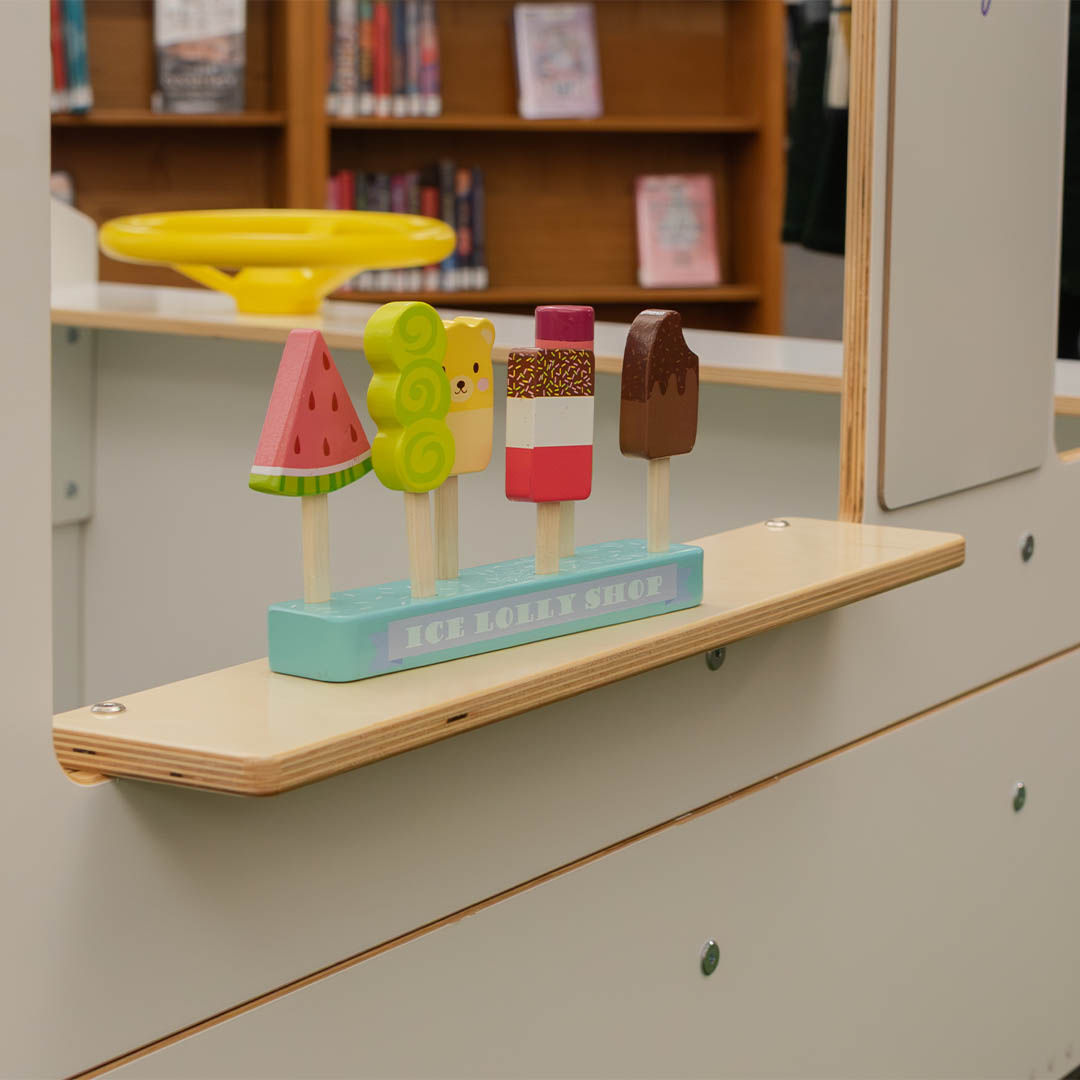 Toy popsicles and ice cream on a wooden stand labeled Ice Lolly Shop, sitting on a wooden ledge attached to a play food truck with a yellow steering wheel and a bookshelf in the background.